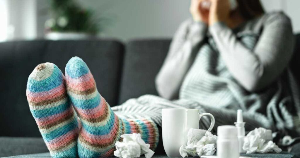 Sick woman with flu, cold, fever and cough sitting on couch at home. Ill person blowing nose and sneezing with tissue and handkerchief. Woolen socks and medicine. Infection in winter. Resting on sofa.