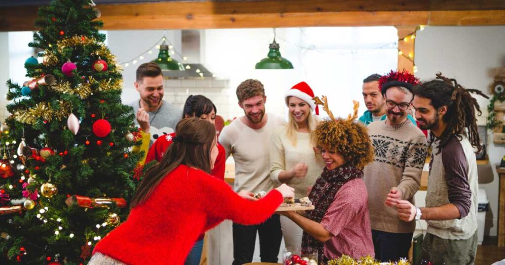 A group of friends are decorating the Christmas tree together. A woman is holding some cake and all the friends are digging in and enjoying themselves.